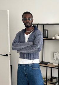 Strikket blå cropped sweater med lodrette striber, båret over en hvid T-shirt, kombineret med løse blå jeans. Klare solbriller og kæde-detaljer.