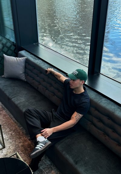 Sofá tapizado oscuro con detalle acolchado, almohada gris, persona vestida con una camiseta negra y una gorra verde, zapatos a rayas grises, gran ventana de vidrio.