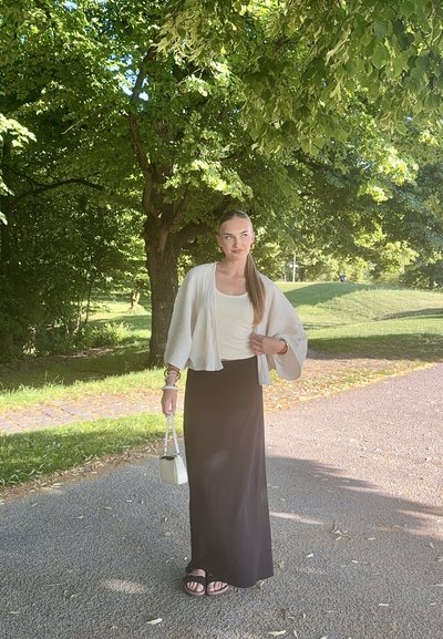 Cárdigan gris claro, blusa blanca, falda larga negra, sandalias negras. Lleva un pequeño bolso blanco; entorno al aire libre con vegetación de fondo.