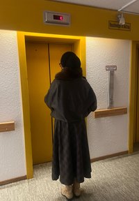 Výtah se žlutými dveřmi, osvětleným tlačítkem s písmenem „E“ a dřevěným madlem v blízkosti. Osoba v tmavém kabátu a vzorované sukni čeká.