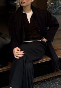 Manteau en laine noire porté sur un cardigan marron, un haut blanc et un pantalon à fines rayures. Sac en cuir noir et ongles manucurés visibles. Banc en bois en arrière-plan.