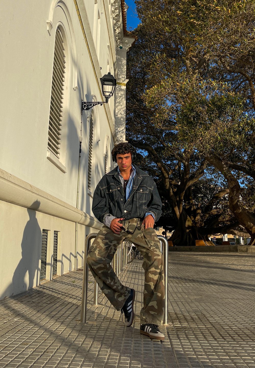 Chaqueta de mezclilla sobre una camisa azul, pantalones de camuflaje, zapatillas negras y blancas, sentado en un riel de metal con un fondo de pavimento texturizado.