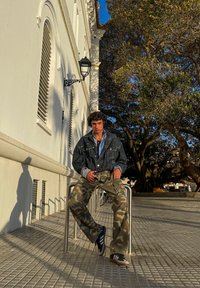Chaqueta de mezclilla sobre una camisa azul, pantalones de camuflaje, zapatillas negras y blancas, sentado en un riel de metal con un fondo de pavimento texturizado.