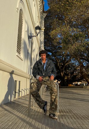 Chaqueta de mezclilla sobre una camisa azul, pantalones de camuflaje, zapatillas negras y blancas, sentado en un riel de metal con un fondo de pavimento texturizado.
