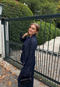Dunkelblaue Jeansjacke mit Kragen und Taillenzug, kombiniert mit weit geschnittenen Hosen. Hält eine kleine schwarze Handtasche. Hintergrund aus grünen Pflanzen und Kies.