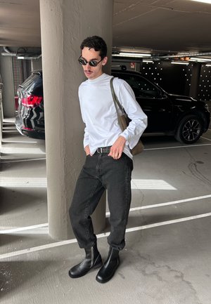 Hombre con camisa blanca de manga larga, jeans negros y botas, de pie en un estacionamiento junto a un pilar de concreto, con gafas de sol y bolso al hombro.