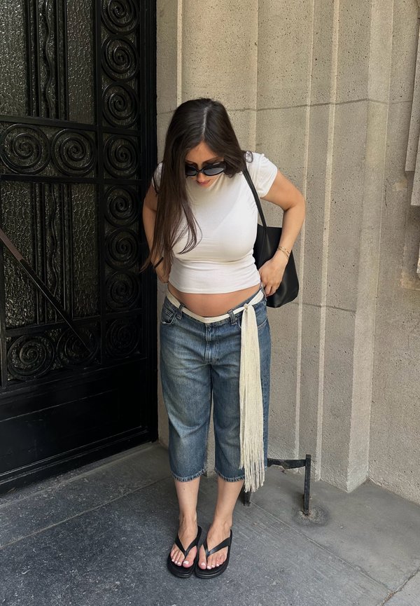 Femme portant des lunettes de soleil, un top court blanc, un jean ample avec une ceinture longue à franges et des sandales noires, debout près d'un mur en pierre et d'une porte en fer forgé noire.