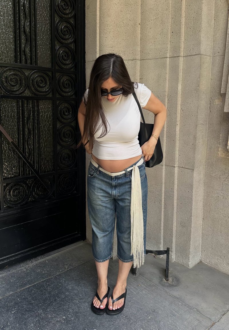 Femme portant des lunettes de soleil, un top court blanc, un jean ample avec une ceinture longue à franges et des sandales noires, debout près d'un mur en pierre et d'une porte en fer forgé noire.