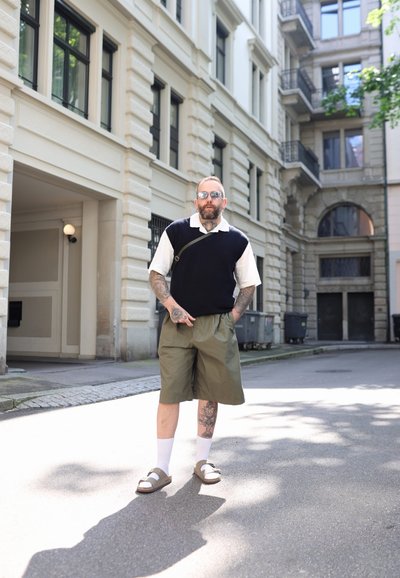 Hombre con un suéter negro sin mangas sobre una camisa blanca, pantalones cortos verde oliva y sandalias marrones, de pie en una calle de la ciudad.
