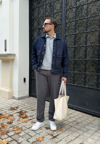 Mężczyzna z brodą i okularami przeciwsłonecznymi, ubrany w dżinsową kurtkę, szare spodnie i białe trampki, stoi na chodniku pokrytym liśćmi, trzymając beżową torbę na ramię.