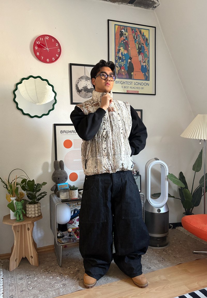 Personne portant des lunettes, un gilet à motifs et un pantalon noir ample, debout pensivement dans un salon avec des plantes, une horloge rouge, des œuvres d'art encadrées et un purificateur d'air moderne.