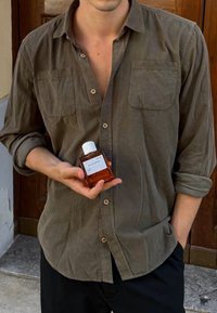 Donker olijfgroene lange mouwen shirt met knoopsluiting aan de voorkant, twee borstzakken, en een glazen parfumfles met een witte dop en label in de hand.