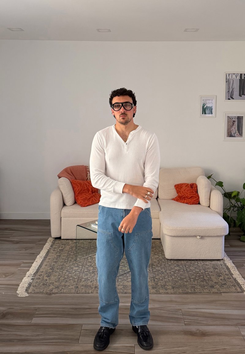 Homem a usar camisa branca de mangas compridas e jeans está numa sala de estar com sofá bege, almofadas laranja, mesa de centro de vidro e arte de parede emoldurada.