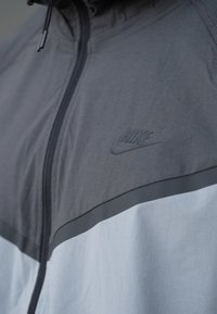 Kurtka Nike w kolorze szarym i jasnoniebieskim, z zamkiem błyskawicznym i miękką fakturą, z subtelnym tłoczonym logo na piersi.