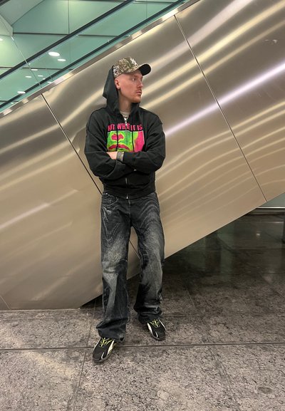 Sudadera negra con un diseño gráfico colorido, jeans azules desgastados y zapatillas negras con acentos verdes, de pie frente a un fondo metálico.