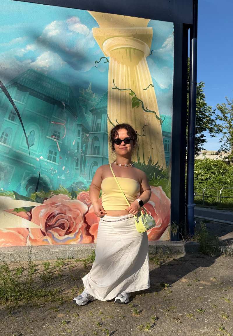 Personne portant des lunettes de soleil, un top jaune, une jupe blanche et un petit sac jaune, debout devant une fresque avec des fleurs et un grand pilier.