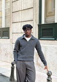 Pull gris texturé en tricot avec un col, associé à un pantalon noir à fines rayures. Béret noir avec un petit logo, sur fond de rue urbaine.