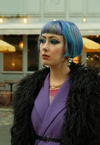 Coiffure bob courte colorée avec des nuances de bleu et de vert. Portant un manteau violet avec de la fausse fourrure noire et un collier en chaîne dorée.