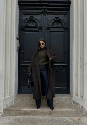 Abrigo oversized marrón oscuro, suéter de cuello alto verde oliva, jeans anchos oscuros, bolso negro y tacones negros puntiagudos frente a una puerta negra.