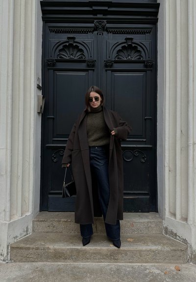Abrigo oversized marrón oscuro, suéter de cuello alto verde oliva, jeans anchos oscuros, bolso negro y tacones negros puntiagudos frente a una puerta negra.