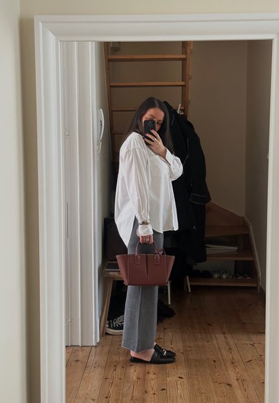 Blusa blanca de manga larga con mangas abullonadas, pantalones grises ajustados y un bolso estructurado marrón, con un reflejo en el espejo de fondo.
