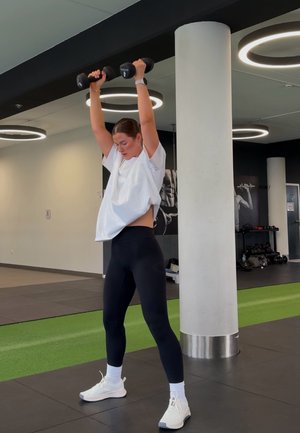 Mujer levantando mancuernas negras por encima de la cabeza en un gimnasio con paredes blancas, luces circulares en el techo y equipo de entrenamiento en el fondo.