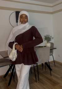 Haut péplum bordeaux avec boutons, hijab blanc et sac à perles ; pantalon large crème ; intérieur minimaliste avec des chaises et des plantes.
