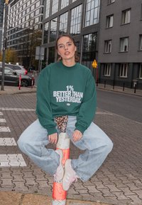 Zelená mikina s bílým textem "1% LEPŠÍ NEŽ VČERA", v kombinaci s volnými světle modrými džínami a růžovými teniskami, sedící na sloupu.