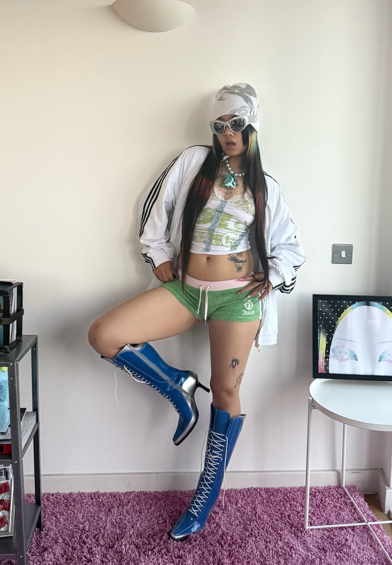 Personne portant des bottes bleues à talons hauts à lacets, un short vert, une veste blanche, des lunettes de soleil et un foulard, posant avec une jambe levée à l'intérieur sur un tapis violet.