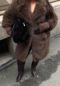 Brun fuskpälsjacka med bred krage, ribbade bruna ärmar, svart axelväska, eleganta spetsiga bruna stövlar. Mjuka texturer och rika färger.