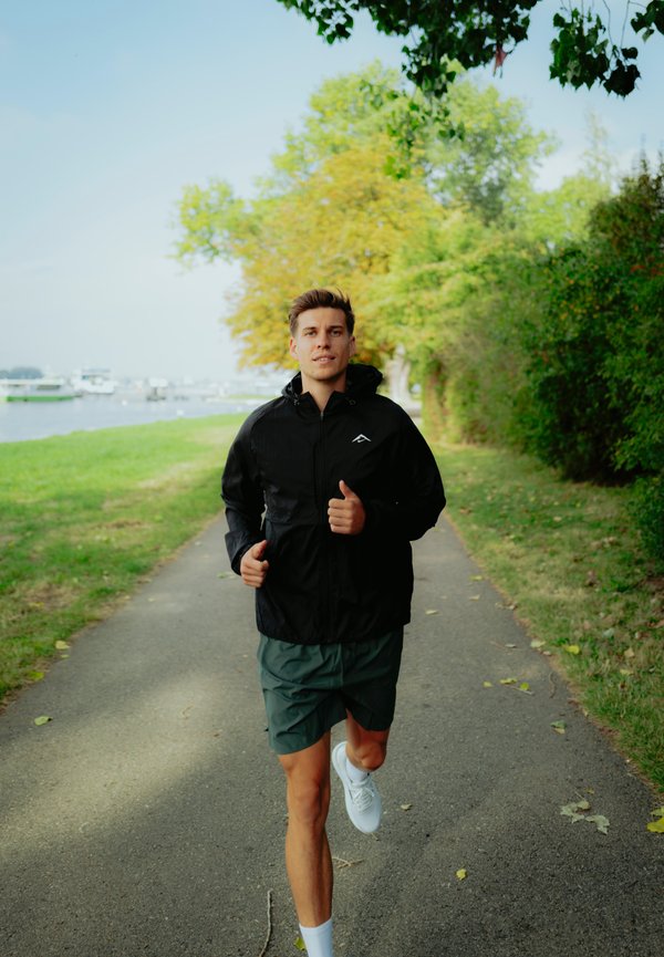 Chaqueta de running negra con capucha sobre unos pantalones cortos deportivos verdes. El corredor lleva zapatillas blancas y está trotando por un camino bordeado de árboles junto a un cuerpo de agua.
