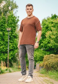 Brun stickad t-shirt med korta ärmar och kontrasterande sömmar, i kombination med slim-fit grå jeans och off-white sneakers, på en väg omgiven av grönska.
