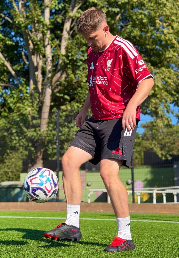Jeune homme en tenue de football de Liverpool jonglant avec un ballon sur un terrain d'herbe avec des arbres et une clôture en arrière-plan.