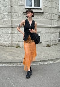 Svart ärmlös väst över en orange spetskjol. Kjolen är genomskinlig med blommönster. Svarta sneakers och en svart axelväska fulländar looken.