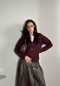 Μπορντό cardigan με κλείσιμο από κουμπιά, συνδυασμένο με καφέ δερμάτινη πλισέ φούστα. Τα αξεσουάρ περιλαμβάνουν μία μικρή καφέ τσάντα και γυαλιά.