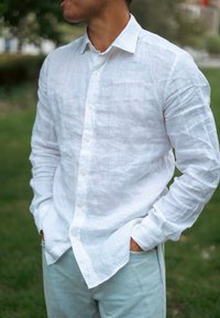 Camisa de linho branca de botão com mangas longas e acabamento texturizado, combinada com jeans azul claro. A camisa está fora da calça, com as mãos nos bolsos.