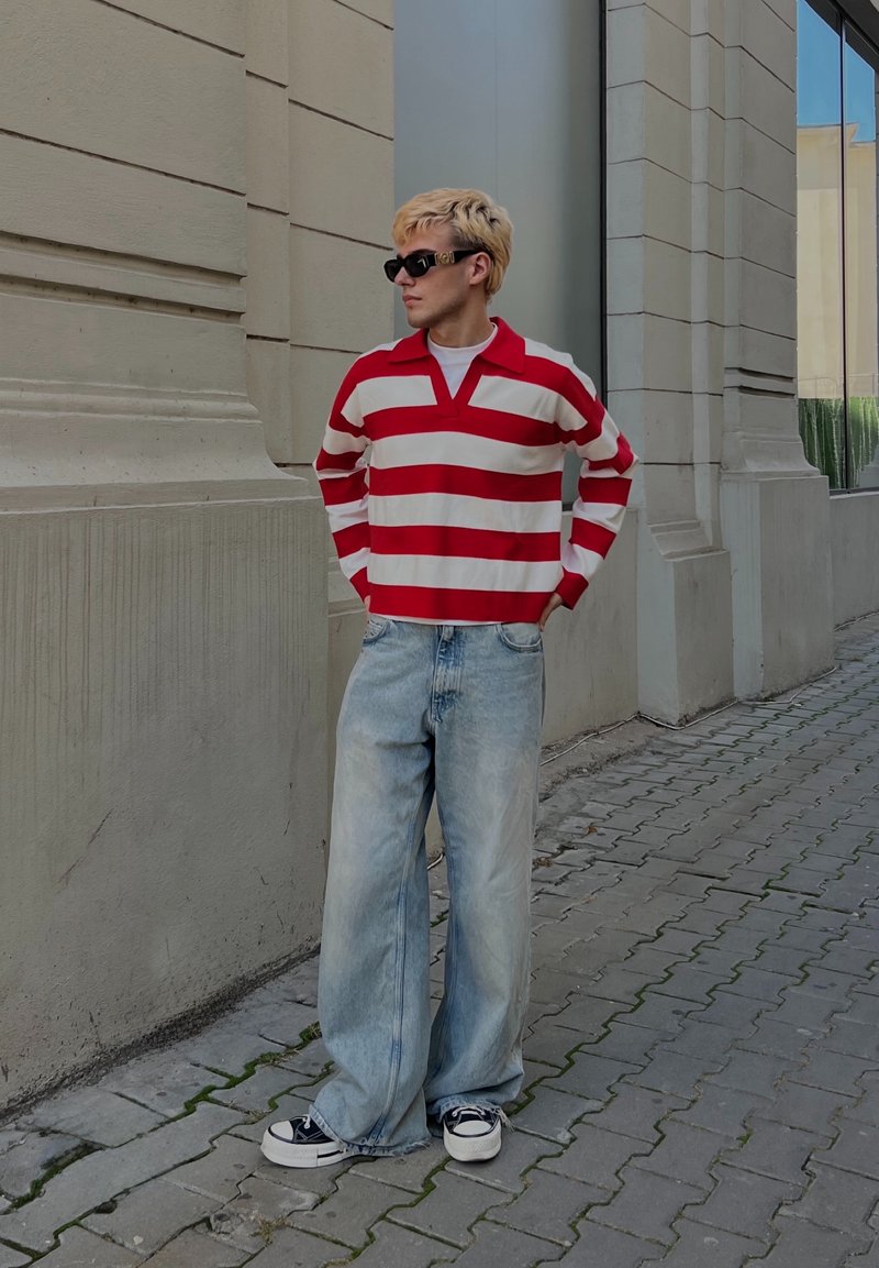 Polo a maniche lunghe a righe rosse e bianche, jeans azzurri a vestibilità comoda, sneakers nere e occhiali da sole. Look casual da strada.