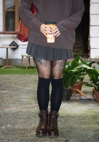 Brun strikket sweater, plisseret sort nederdel, sorte strømpebukser med tatoveringer og brune snørebåndssko. Holder en orange kaffekop.