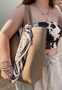 Borsa tote in pelle beige con texture, con una sciarpa annodata a strisce crema e nera attaccata; indossata con un top corto a fantasia e pantaloni chiari.
