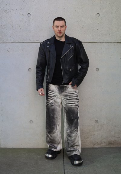 Chaqueta de motociclista de cuero negro con herrajes plateados, camiseta negra ajustada, pantalones gris desgastados y holgados, y zapatos de plataforma negros con acentos.