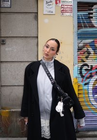 Femme aux cheveux lissés en arrière, vêtue d'un manteau noir, d'un sweat gris à fermeture éclair et d'un sac bandoulière avec un porte-clés peluche blanc, debout près de graffitis et de panneaux.