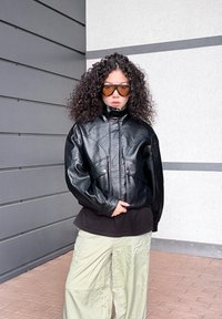 Fekete műbőr dzseki magas nyakkal, két elülső zsebbel és széles ujjakkal, fekete inggel és világos zöld nadrággal párosítva.