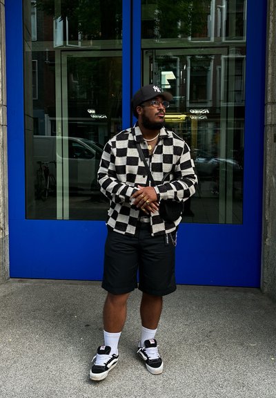 Chaqueta de punto a rayas en blanco y negro, pantalones cortos negros, calcetines blancos y zapatillas negras. De pie frente a una puerta azul.