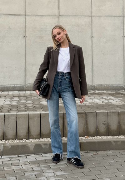 Blazer marrón oscuro sobre una camiseta blanca, combinado con jeans acampanados de color azul claro y zapatillas negras. Sosteniendo un bolso negro con acentos de cadena.