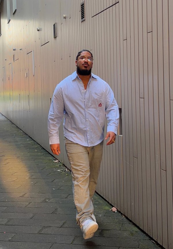 Uomo con capelli intrecciati, occhiali, camicia a righe blu, pantaloni beige e scarpe da ginnastica bianche che cammina lungo un stretto vicolo urbano.