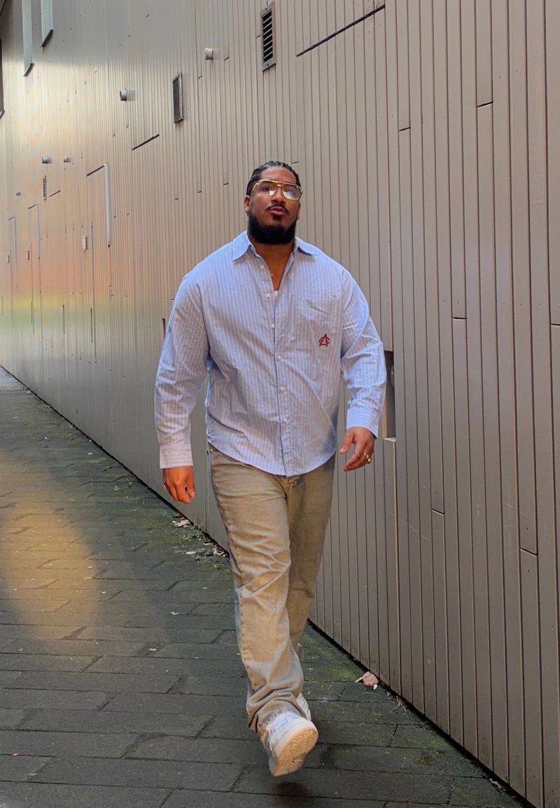 Homme aux cheveux tressés portant des lunettes, une chemise bleue rayée, un pantalon beige et des baskets blanches, marchant le long d'une ruelle urbaine étroite.