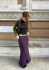 Een aansluitend zwart topje is gecombineerd met een lange paarse rok, voorzien van een gevlochten accentriem. De outfit wordt aangevuld met een groene suede jas.