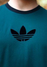 Teal bomull t-skjorte med en svart fløyels Adidas trefoil-logo og tre horisontale striper under. Ribbet svart krage.
