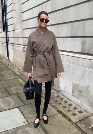 Mujer con abrigo marrón con cinturón, leggings negros y zapatos planos negros, llevando un bolso negro y gafas de sol oscuras en una acera de la ciudad.