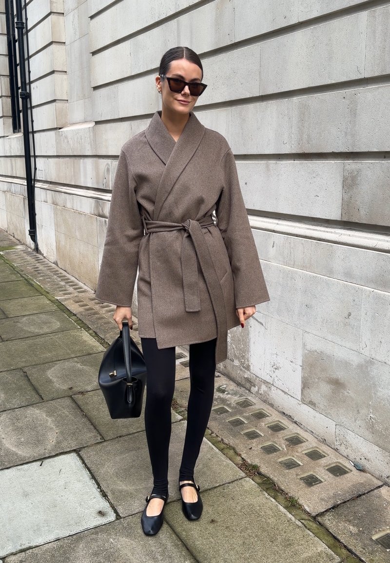 Femme portant un manteau marron ceinturé, des leggings noirs et des ballerines noires, tenant un sac à main noir, avec des lunettes de soleil foncées, sur un trottoir en ville.